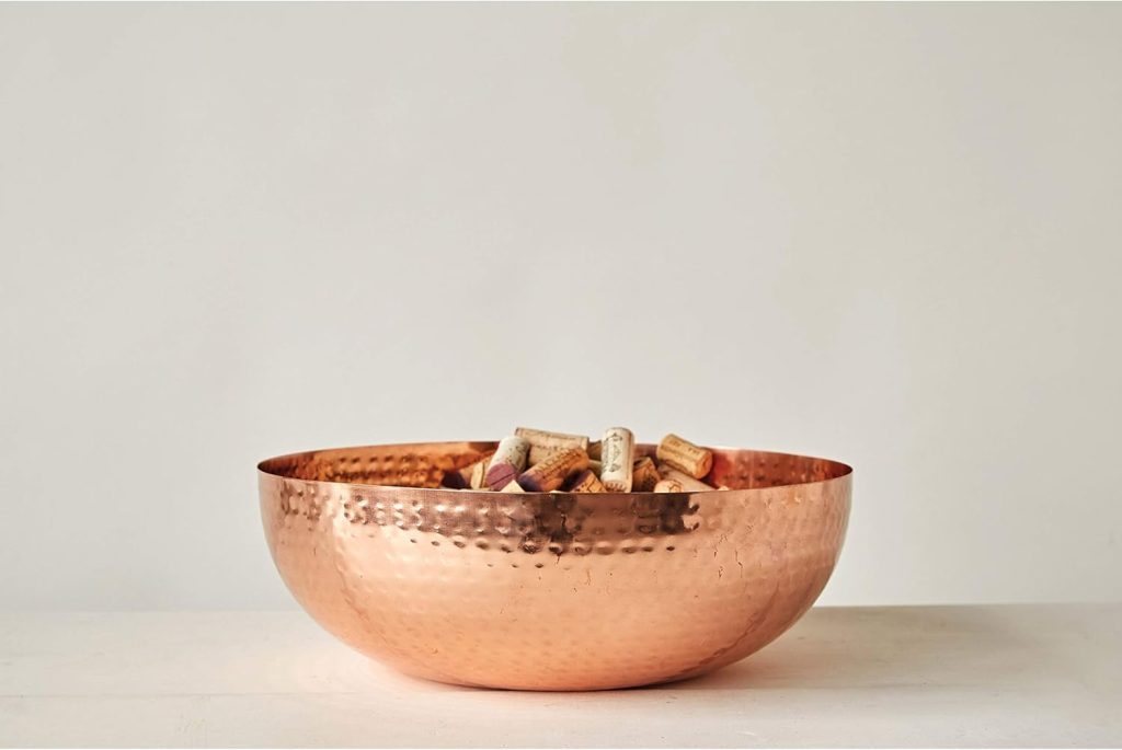 A large hammered copper bowl filled with assorted wine corks sits on a white surface against a plain light background.