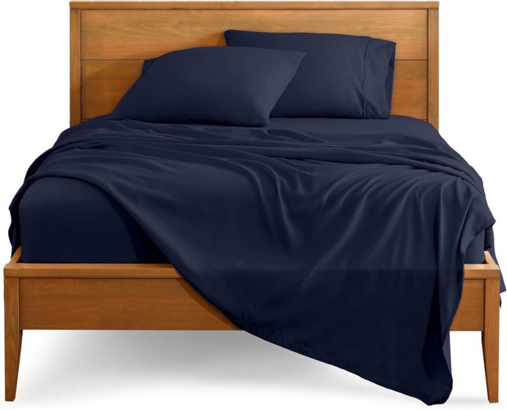 A wooden bed frame with a headboard, made up with dark blue sheets, pillows, and blanket, some bedding slightly draped over the side.