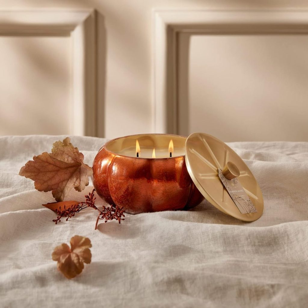 A lit three-wick candle in a copper pumpkin-shaped container sits on a white cloth, with its lid and decorative autumn leaves beside it.