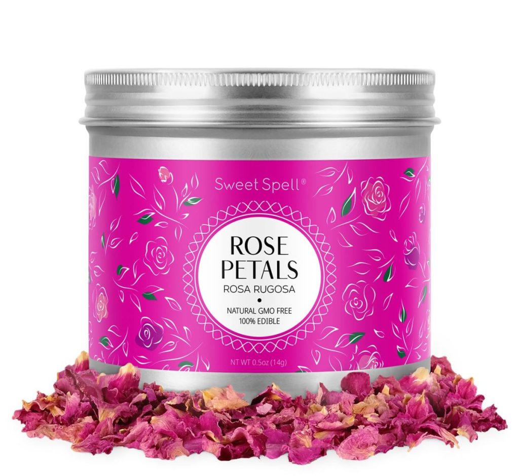 A metal tin labeled "Rose Petals" sits on a pile of dried pink rose petals. The tin has a pink floral design and reads "Sweet Spell, 100% Edible, Natural, GMO Free.