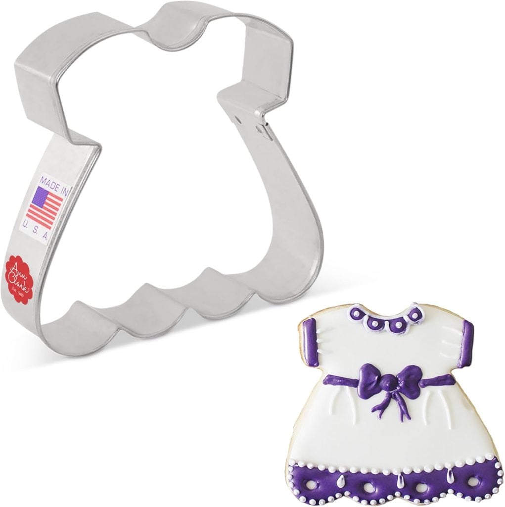 A metal dress-shaped cookie cutter is shown next to a decorated sugar cookie shaped like a dress with white and purple icing details.