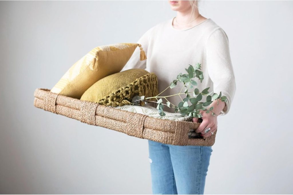 A person in a white top and jeans holds a woven tray containing two yellow cushions and a sprig of greenery.