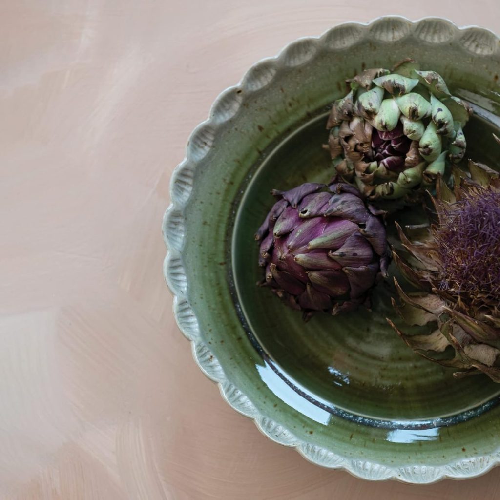 Three artichokes, one green, one purple, and one with a purple thistle flower, arranged on a green plate with a scalloped edge, set against a beige background.