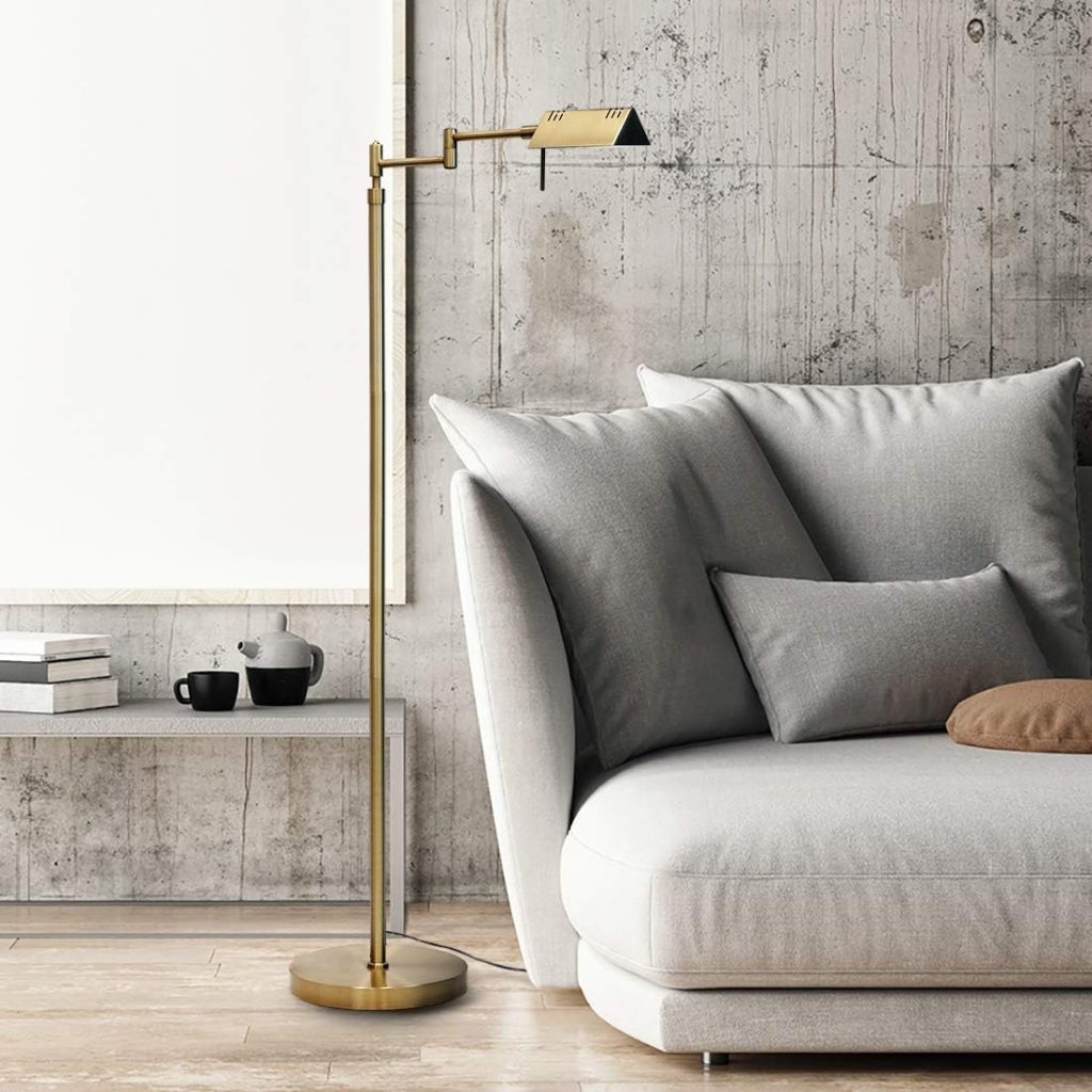 Brass floor lamp stands beside a light gray sectional sofa with pillows; background features a concrete wall and a white shelf with a teapot, cups, and books.