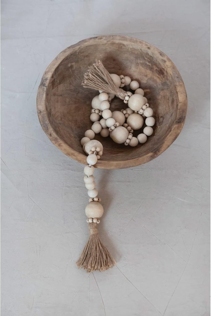 A wooden bowl holds a string of round, natural-colored wooden beads with a twine tassel at the end, resting on a light gray textured surface.