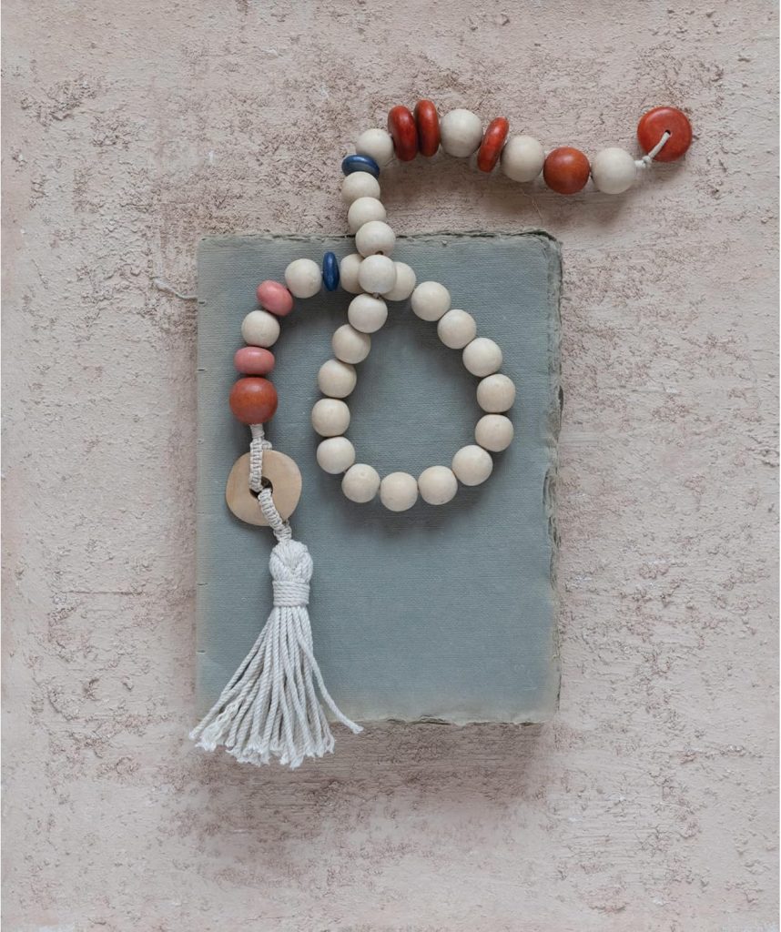 Wooden bead garland with red, blue, and natural beads, a wooden ring, and a white tassel, arranged on a textured stone slab against a pale pink background.