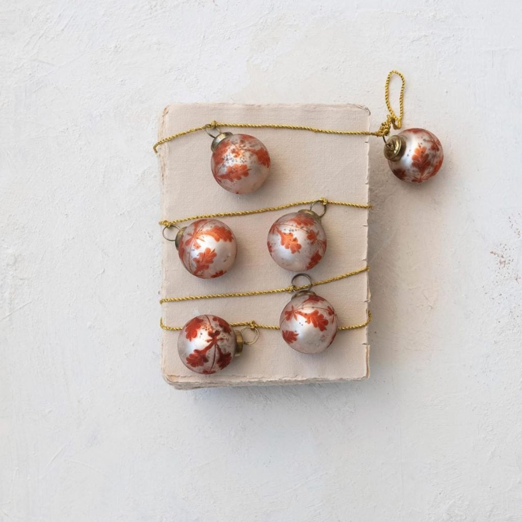 Five round, white Christmas ornaments with red floral patterns are placed on a cream-colored textured surface, four strung on gold thread and one lying separately.
