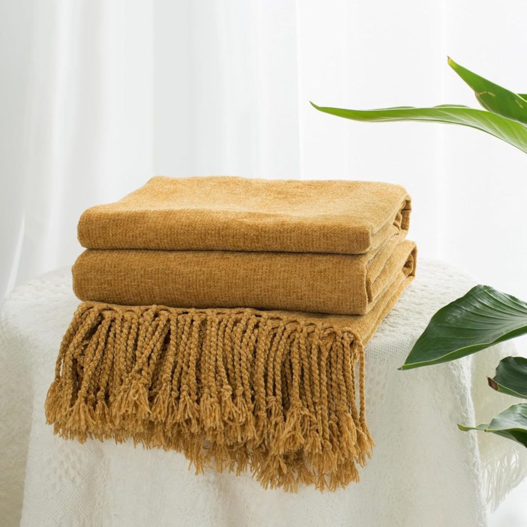 A folded mustard yellow throw blanket with fringed edges sits on a white textured surface next to a green plant.