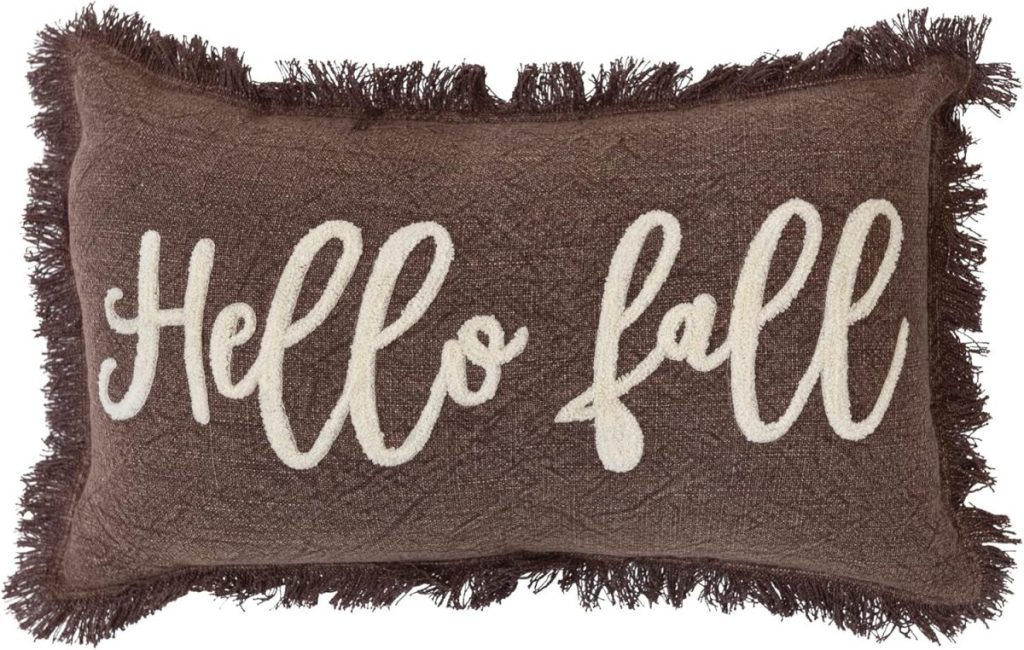 A rectangular brown pillow with fringe edges features the words "Hello fall" in large, white cursive letters.