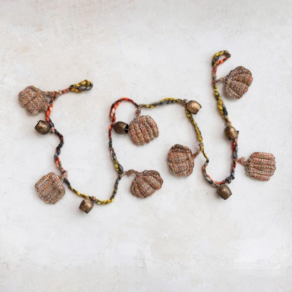 A decorative garland with small, textured pumpkin shapes and metallic bells, connected by a multicolored braided string, displayed on a light surface.