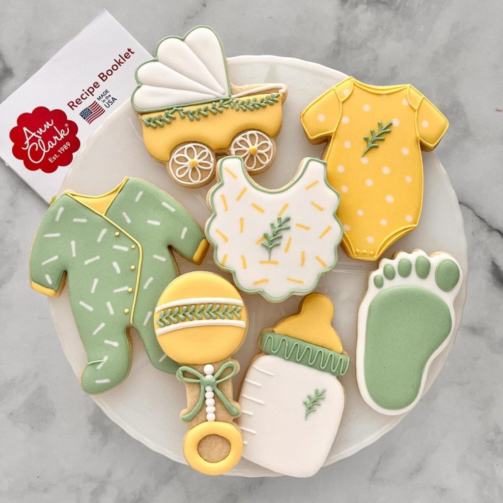 Decorated sugar cookies shaped like a baby bottle, rattle, foot, stroller, onesies, and bib in yellow, green, and white, arranged on a round white tray.
