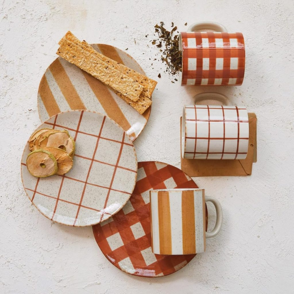 Three ceramic plates and three mugs with various striped and checkered patterns in white and rust tones, displayed on a white surface with dried apple slices and tea leaves.
