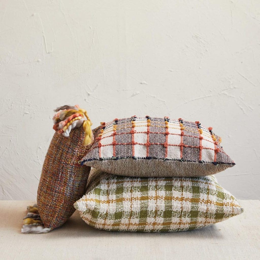 Three textured throw pillows in different plaid and check patterns are stacked against a plain, off-white wall.