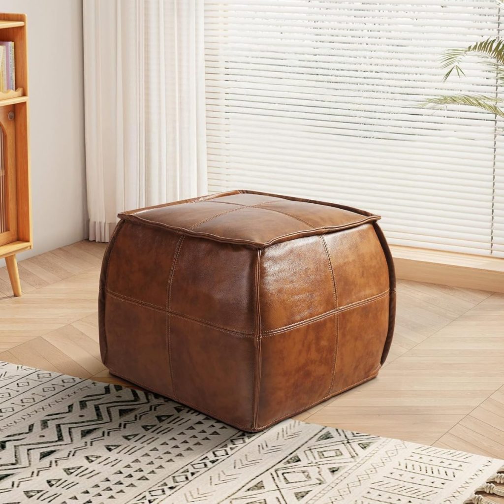 A brown leather pouf sits on a patterned rug in a living room with wooden floors, sheer curtains, and a bookshelf in the background.