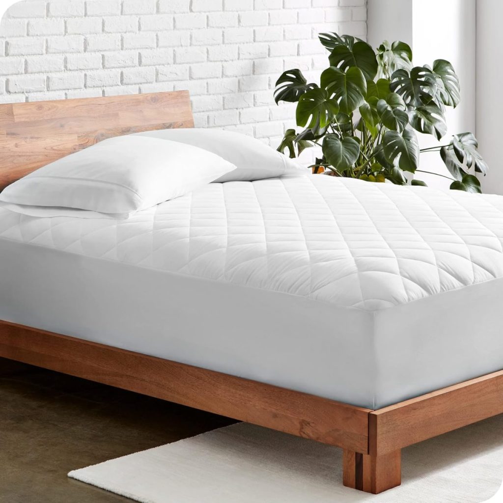 A neatly made bed with a wooden frame, white quilted mattress cover, two white pillows, a white rug, and a large green houseplant in the background.