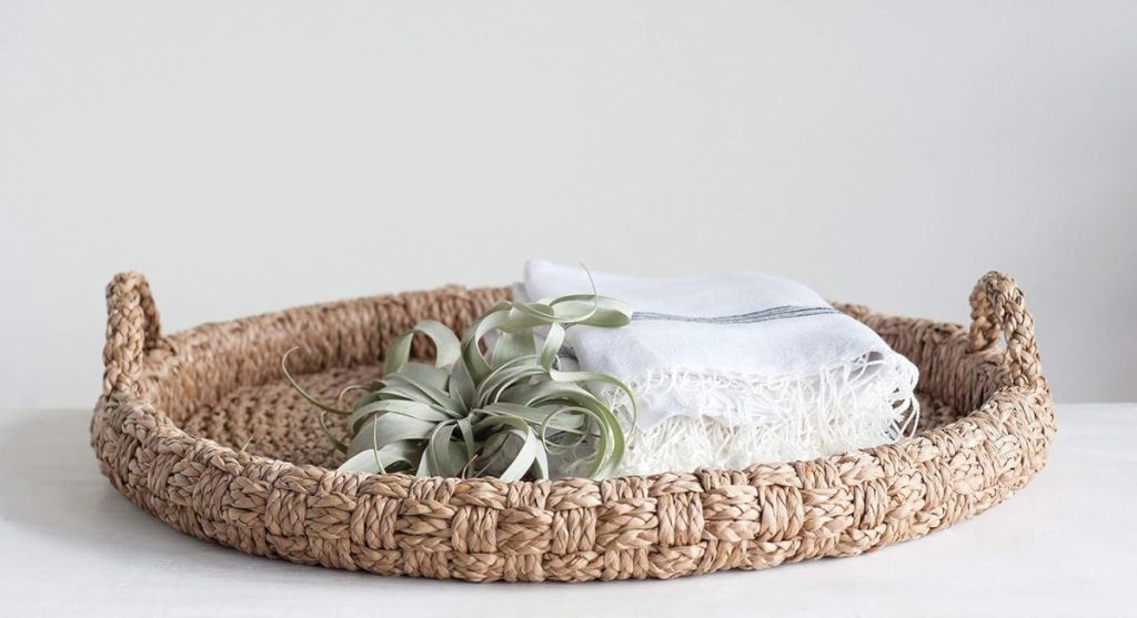 A round woven basket on a white surface holds a folded white fringed cloth and an air plant.