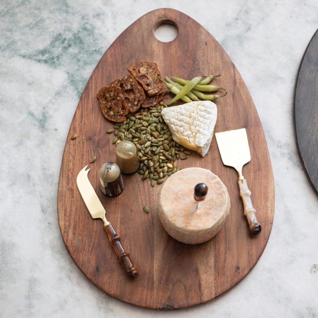 A wooden serving board with crackers, green beans, seeds, brie cheese, a round cheese, salt and pepper shakers, and two cheese knives arranged neatly.