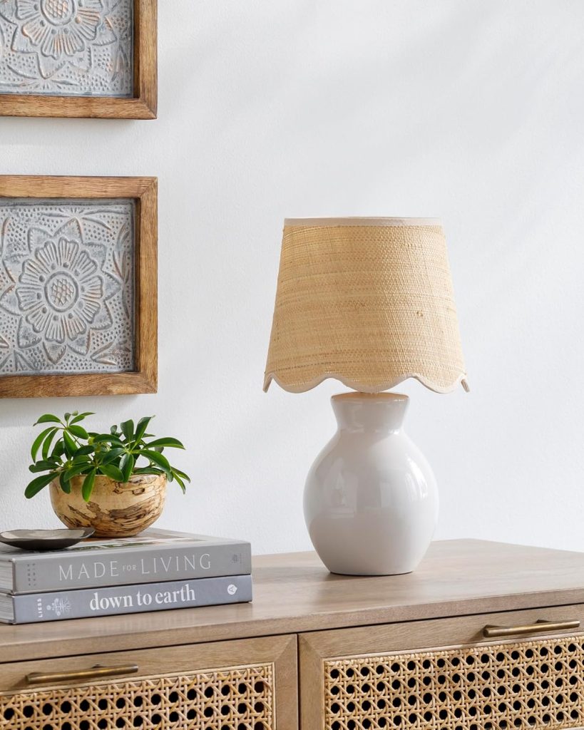 A white ceramic lamp with a woven shade sits on a wooden cabinet beside a small potted plant and two stacked books.