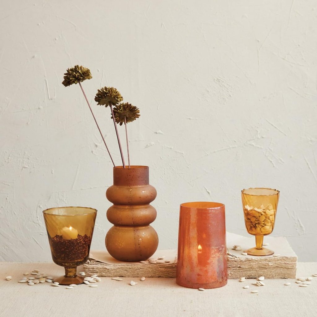 A textured brown vase with dried flowers, two amber goblets, and a translucent orange candle holder are arranged on a beige surface against a light textured wall.