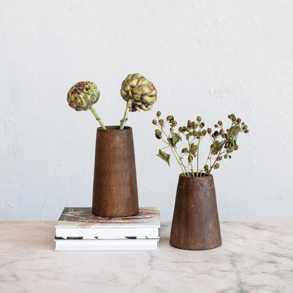 Two wooden vases with dried flowers; one vase sits on a stack of books, the other directly on a marble surface against a plain light background.