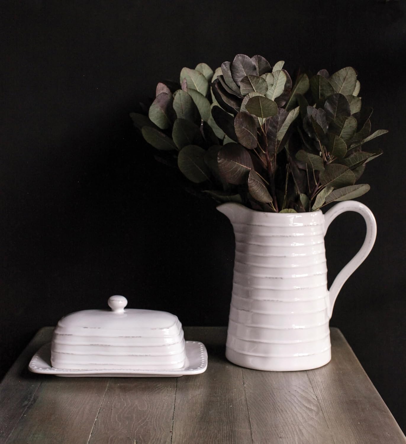 A white ceramic butter dish with a lid sits next to a white ceramic pitcher holding leafy branches on a wooden table against a dark background.