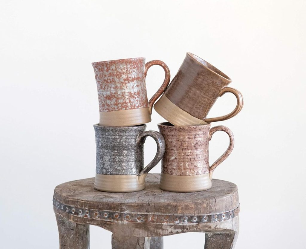 Four ceramic mugs in different earth tones are stacked in pairs on a rustic wooden stool against a plain light background.