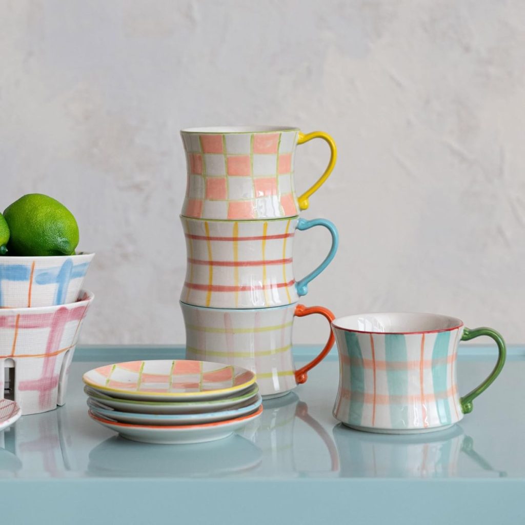 Four plaid-patterned mugs, three stacked and one beside a stack of matching saucers, sit on a light blue surface next to a bowl of limes.