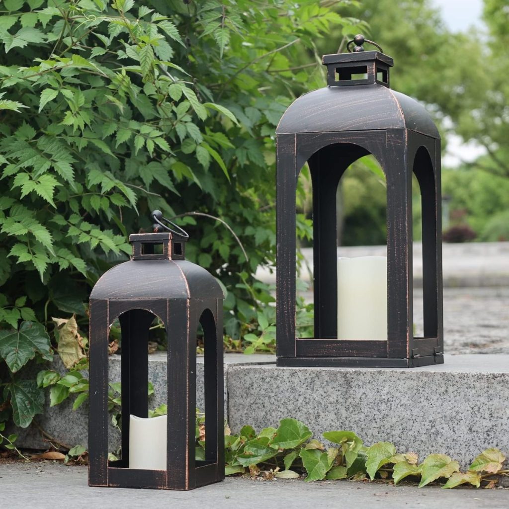 Two black metal lanterns with white pillar candles inside placed on concrete steps next to green leafy plants.