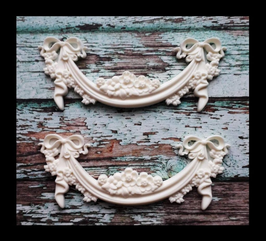 Two ornate white decorative appliques with floral and ribbon designs are elegantly placed beside a ceramic planter on a distressed, weathered teal wooden surface.