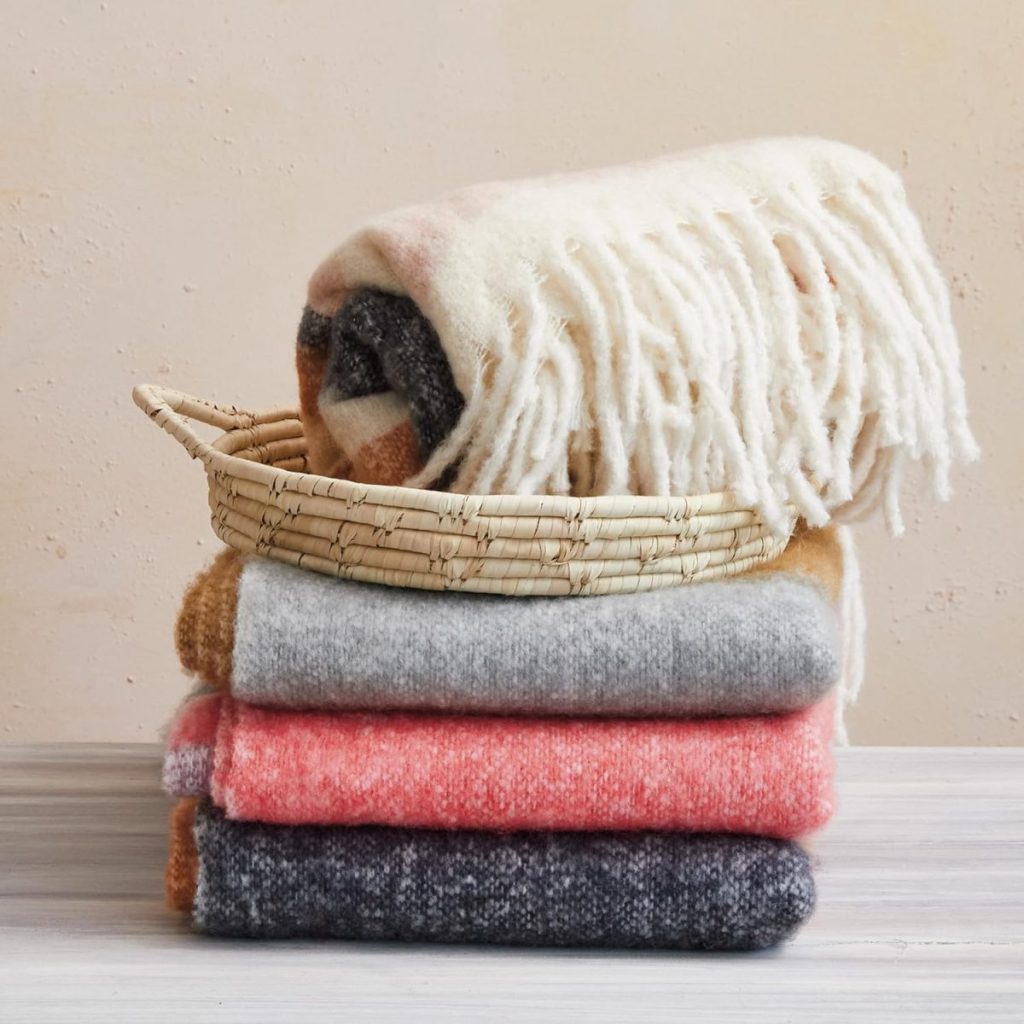 A woven basket holds a fringed, rolled blanket placed on top of a neatly stacked pile of three folded blankets in various colors on a light wooden surface.