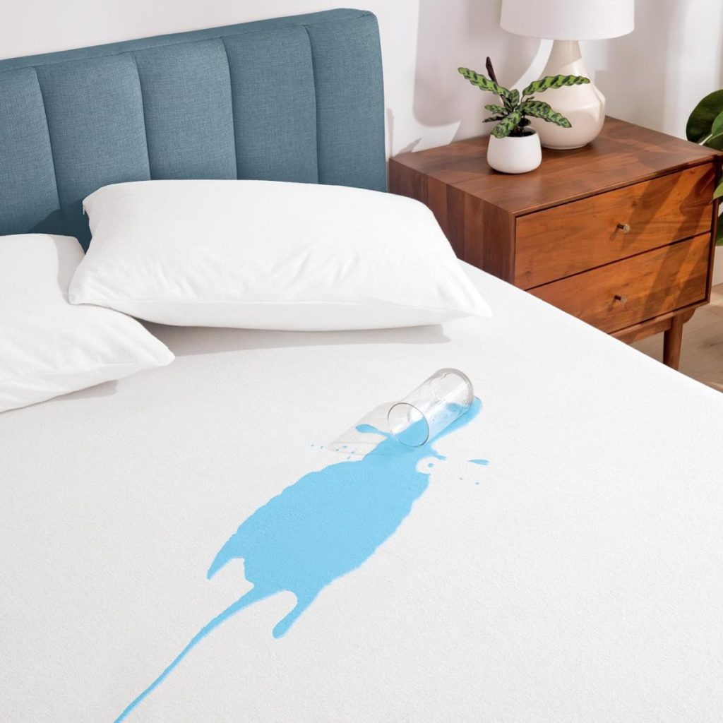 A clear glass tipped over, spilling blue liquid onto a white mattress with white bedding, next to a wooden nightstand and lamp.