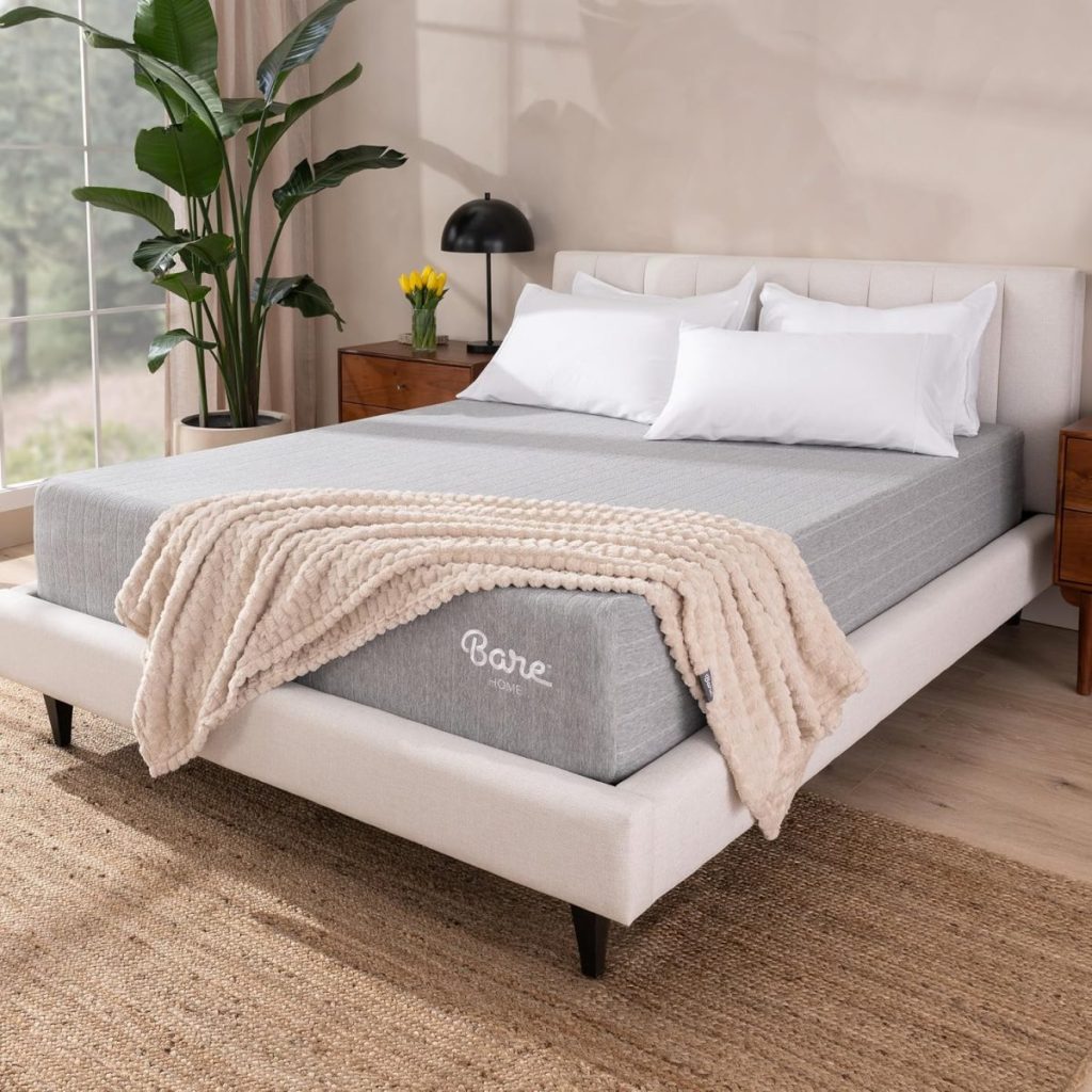 A modern bedroom with a light upholstered bed, gray Bare mattress, white bedding, a beige knit blanket, wooden nightstands, a lamp, potted plant, and a window with natural light.