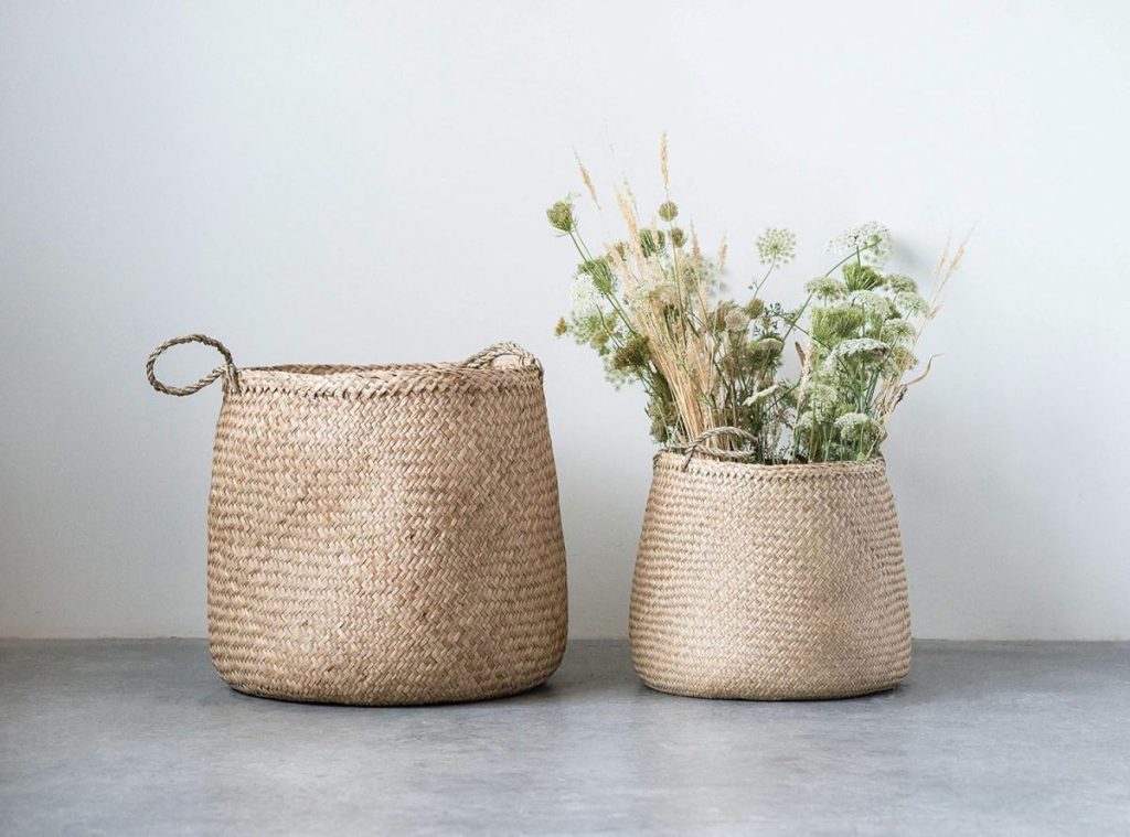 Two woven straw baskets sit on a grey surface; the larger basket is empty, while the smaller basket holds dried grasses and white flowers.