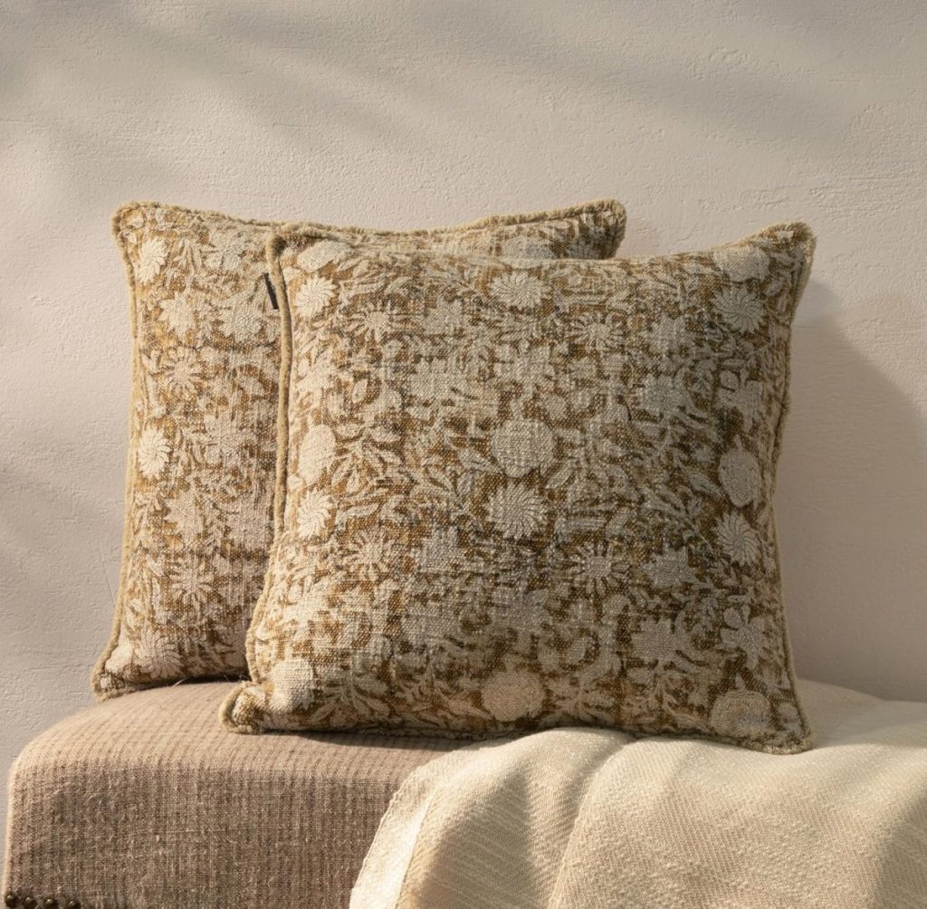 Two square decorative pillows with a beige and brown floral pattern rest against a textured light-colored wall on a neutral woven surface.