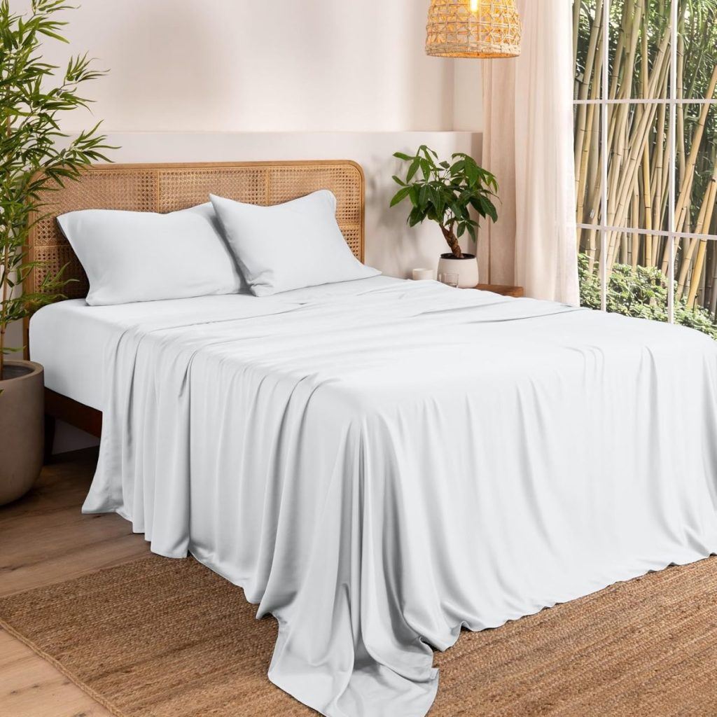 A neatly made bed with white bedding, two pillows, wooden headboard, potted plants, woven rug, and bamboo seen through a window in a bright, modern bedroom.