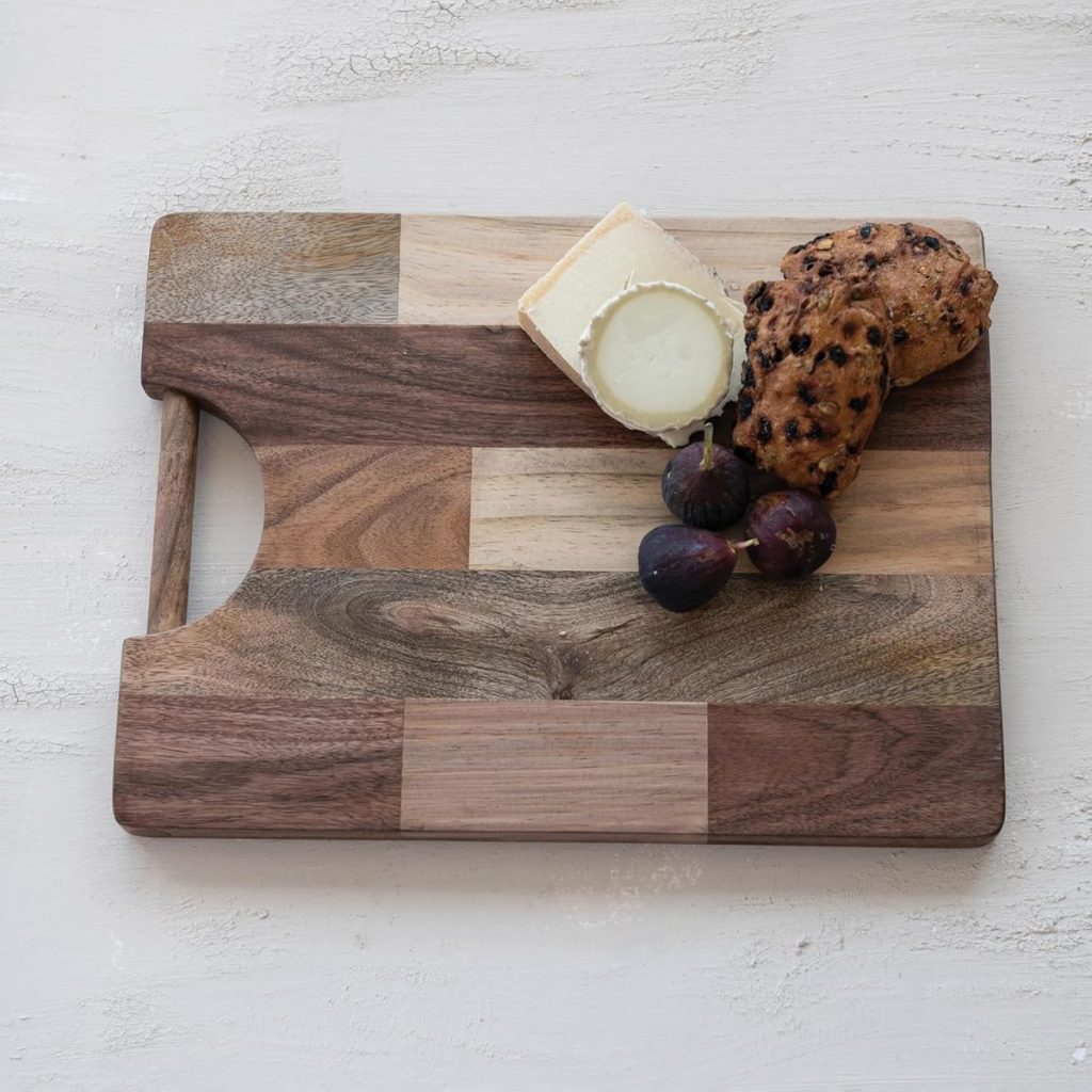 A wooden cutting board with a handle holds a wedge of cheese, two figs, and a chunk of bread with raisins, set on a light textured surface.