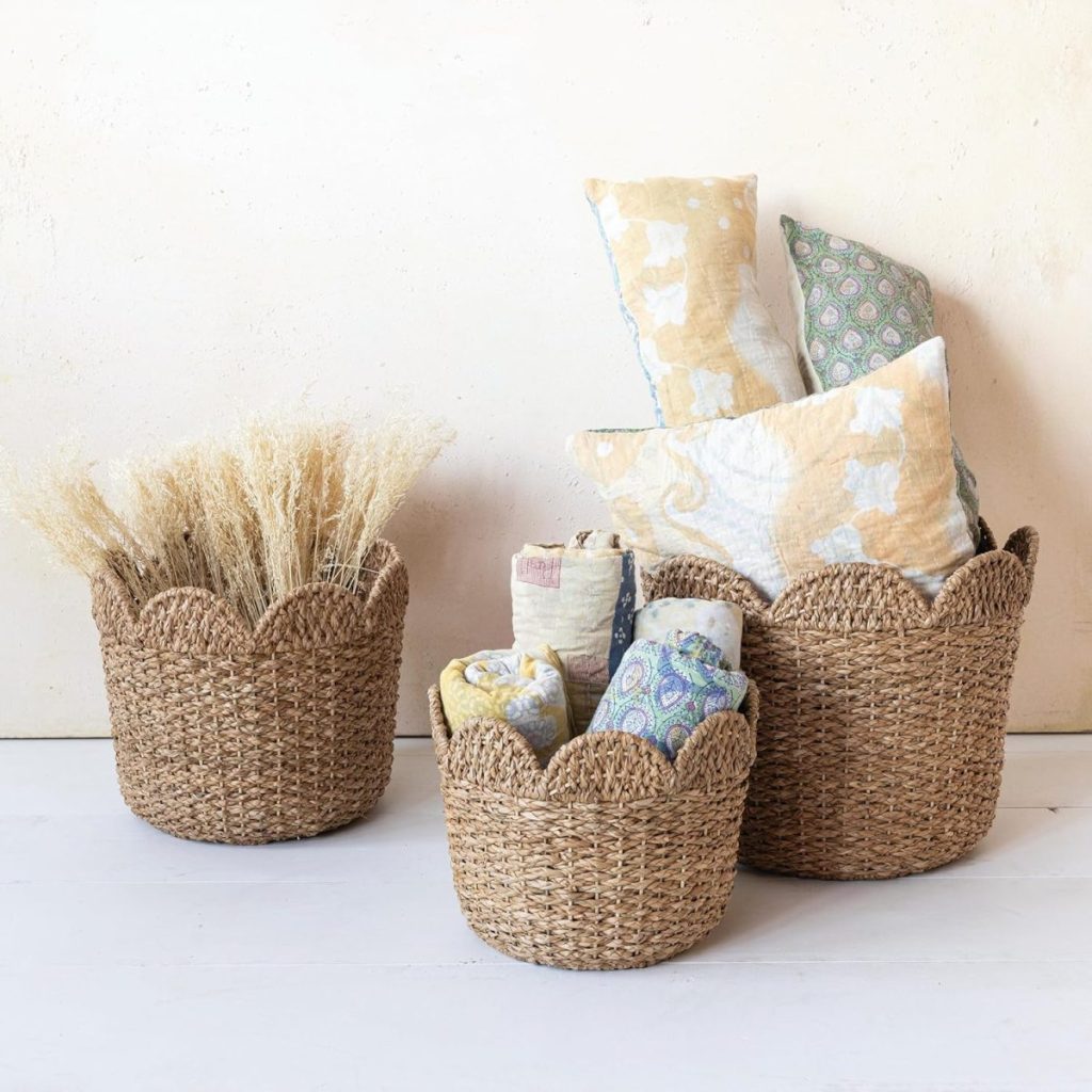 Three woven baskets are shown; one holds dried grasses, another contains rolled fabrics, and the largest contains patterned pillows and textiles against a plain background.