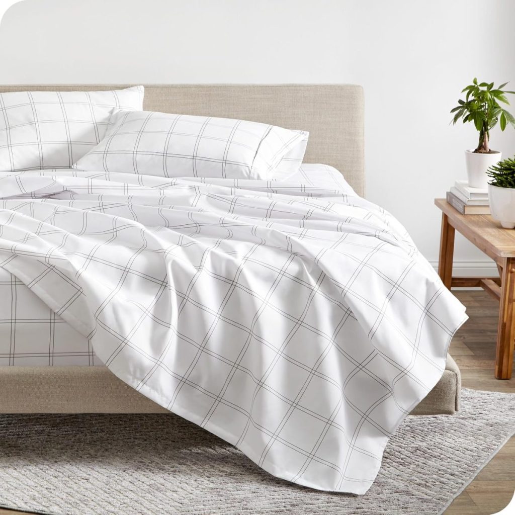 A neatly made bed with white sheets and pillowcases featuring a grey check pattern, next to a wooden nightstand with potted plants and books.