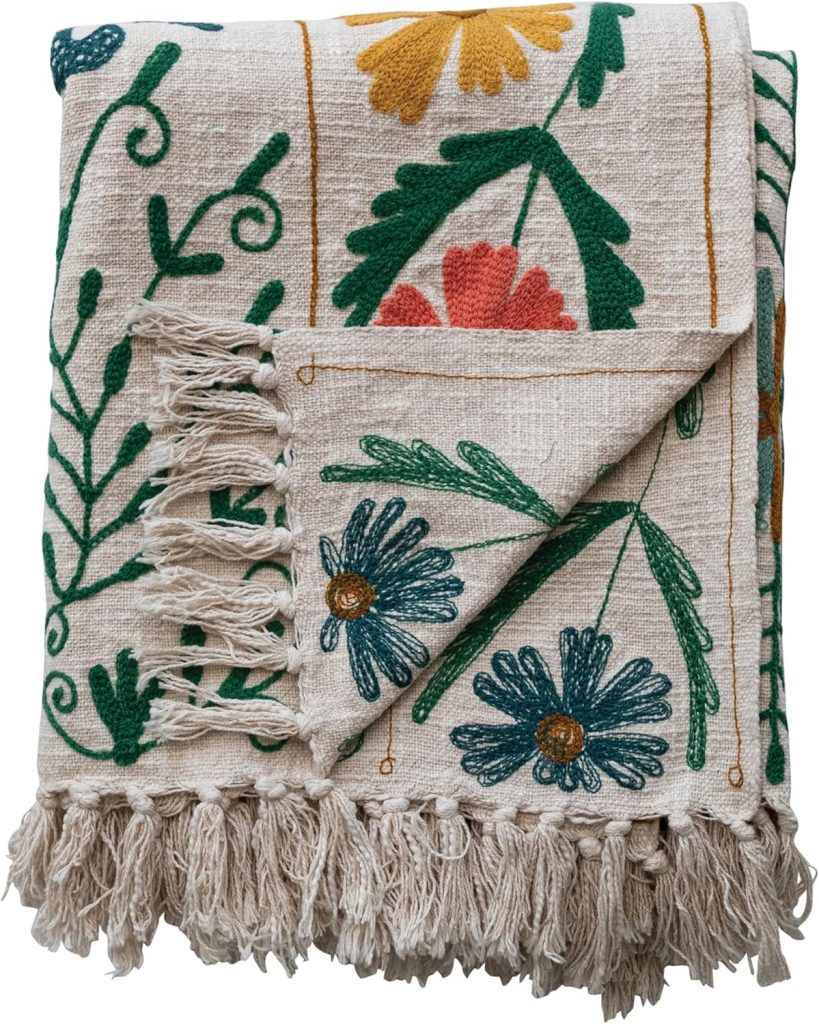 Folded beige blanket with colorful floral embroidery and fringed tassels along one edge.