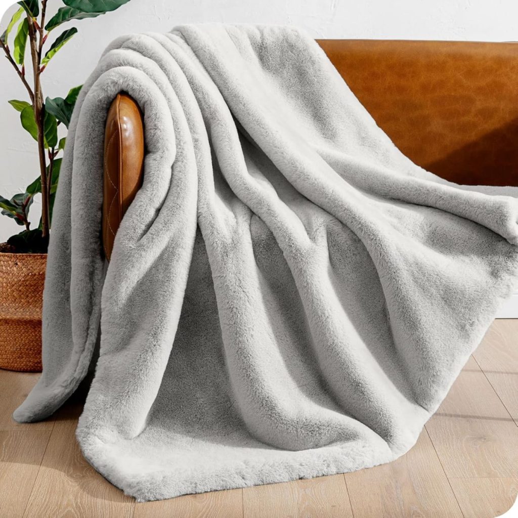 A light gray fluffy blanket draped over the arm of a brown leather sofa, with a potted plant nearby on a wooden floor.