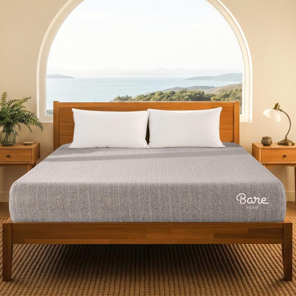 A neatly made bed with a gray mattress, three white pillows, and wooden frame sits in a room with a large arched window overlooking water and greenery.