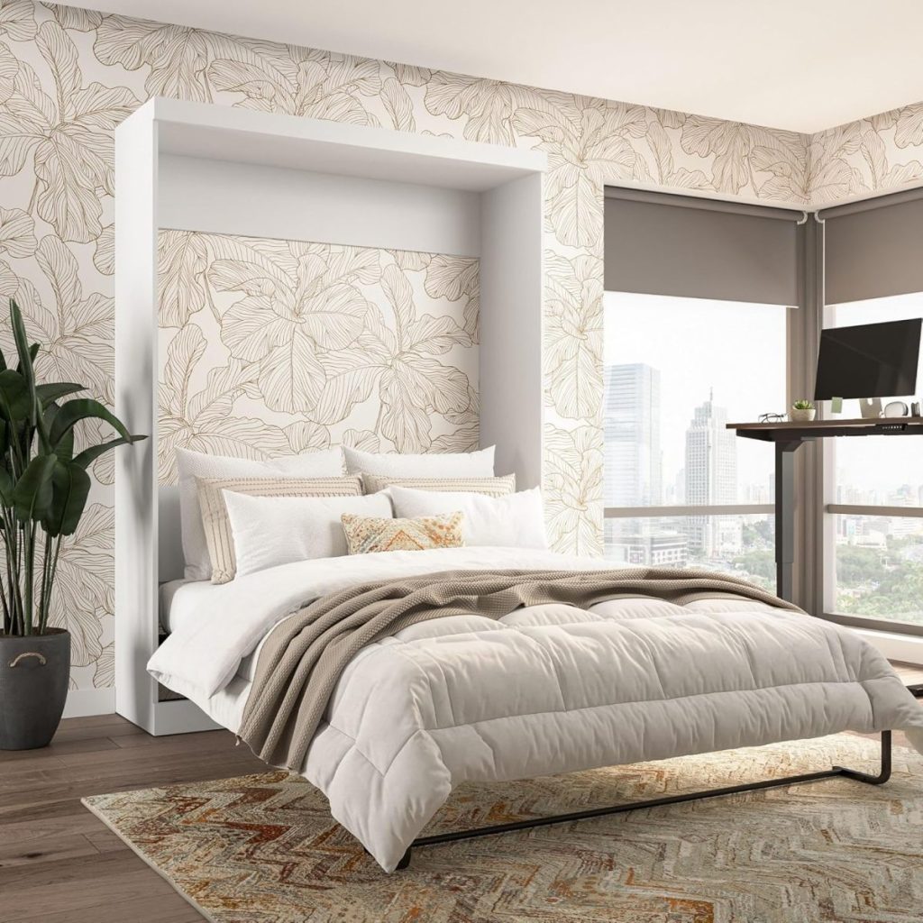 A neatly made bed with beige bedding sits in a modern bedroom with floral wallpaper, large windows, an indoor plant, and a desk with a computer.