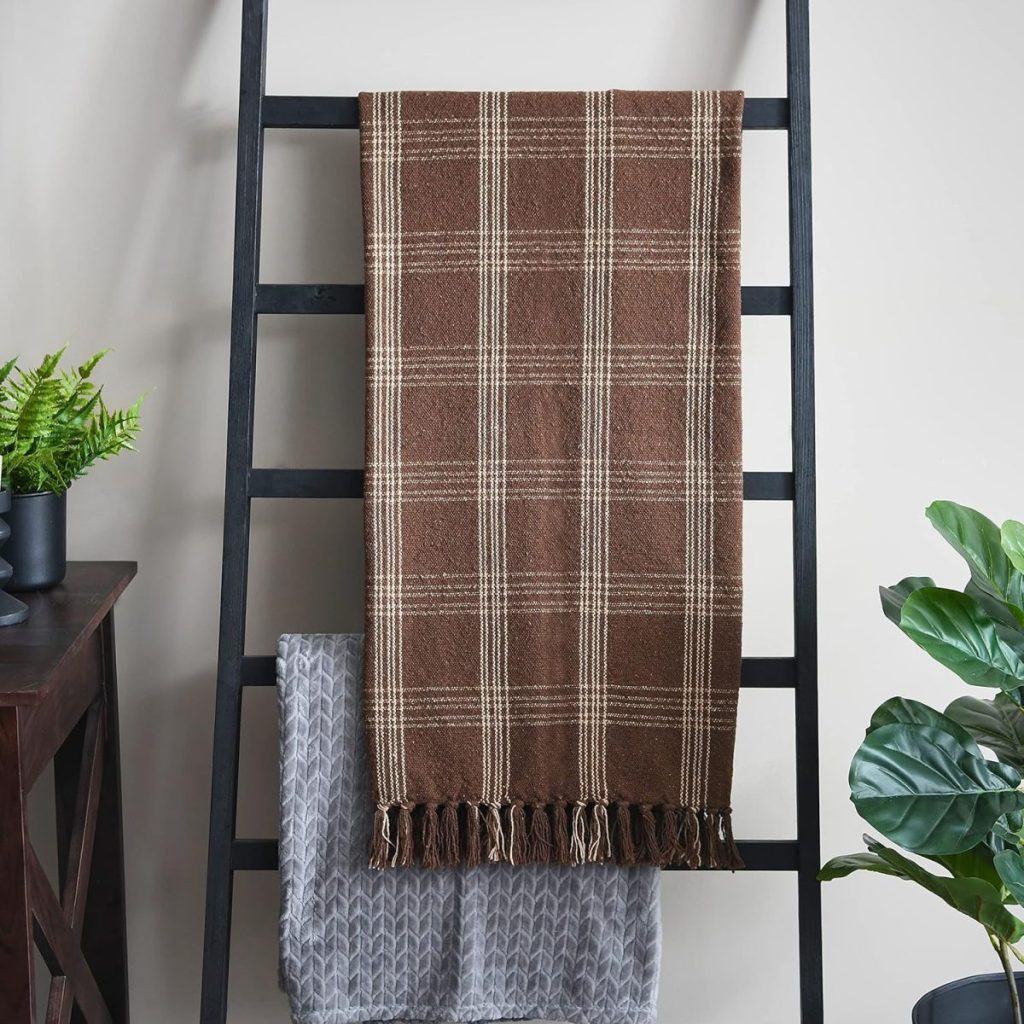 A brown plaid throw blanket with fringe hangs on a black ladder rack above a gray textured blanket, with green potted plants nearby.