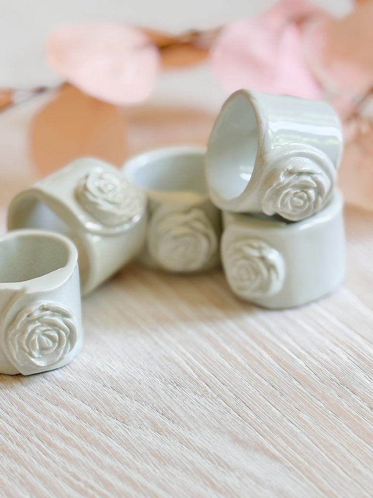 Six white ceramic napkin rings with floral rose designs are stacked on a light wood surface, with pink leaves in the background.