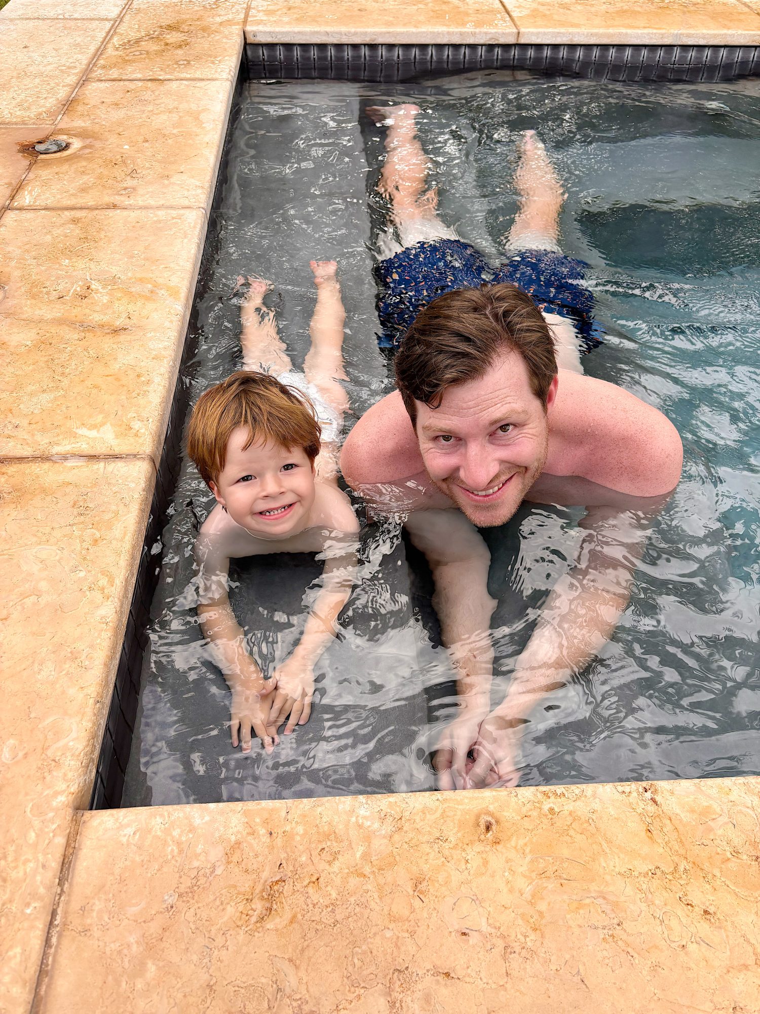 An adult and a child are lying in shallow pool water, smiling at the camera near the edge of a tiled pool surrounded by tan stone.