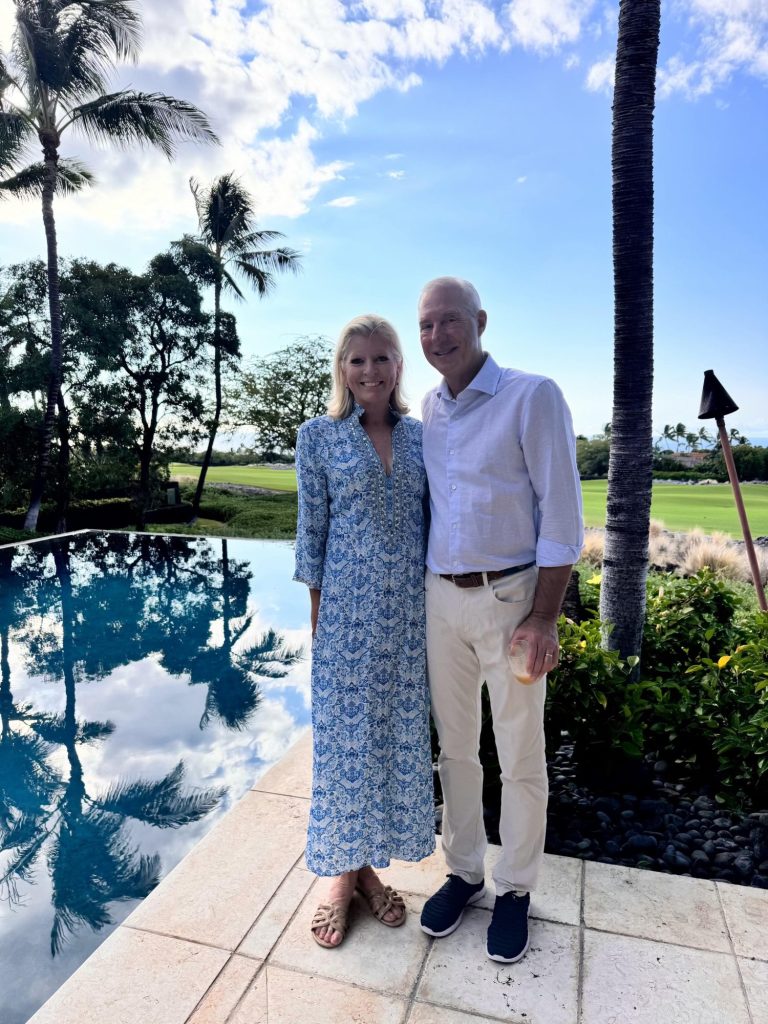 A woman and a man stand side by side by a pool, with palm trees, greenery, and a blue sky with clouds in the background.