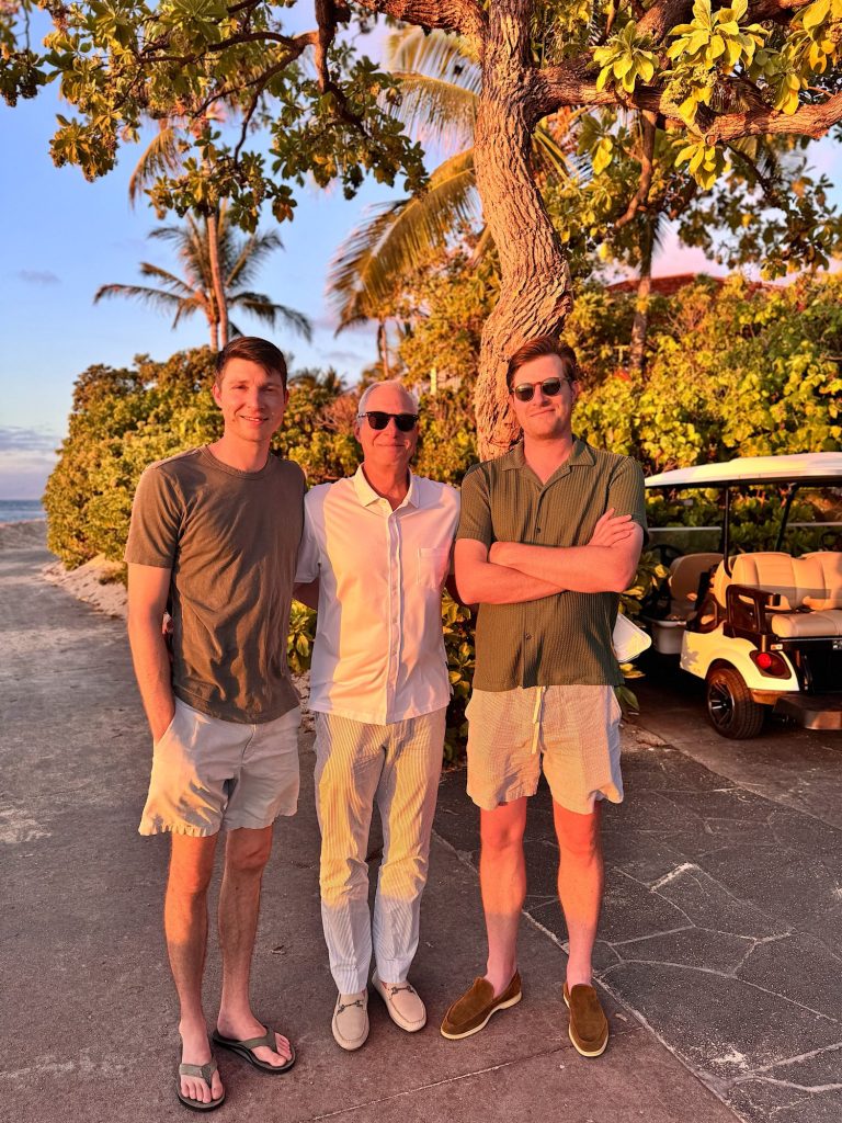 Three men stand together on a sunlit path near a golf cart and palm trees, dressed in casual summer clothing.