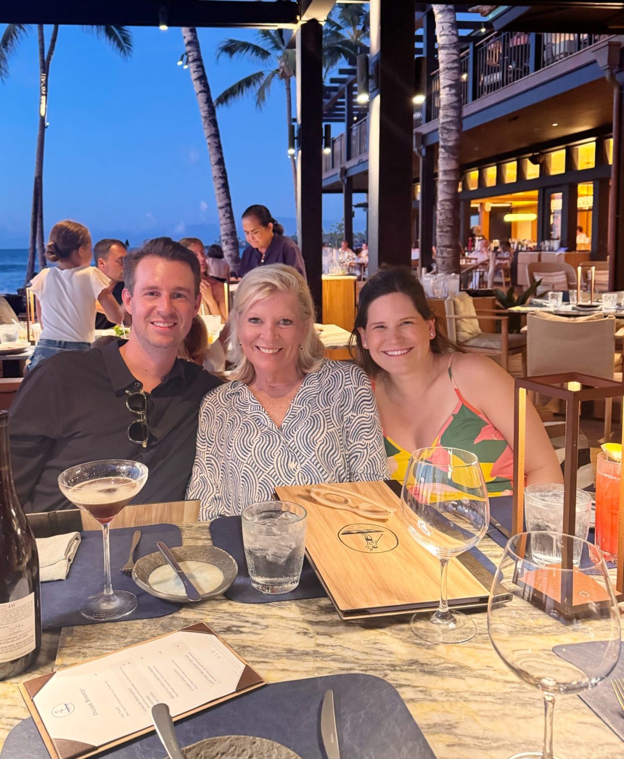 Three people sit together at an outdoor restaurant table in the evening, smiling at the camera. Palm trees and diners are visible in the background.