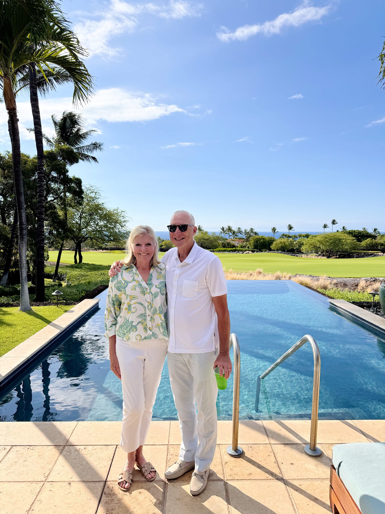 A man and woman stand side by side by a rectangular pool, with palm trees, grass, and a blue sky in the background.