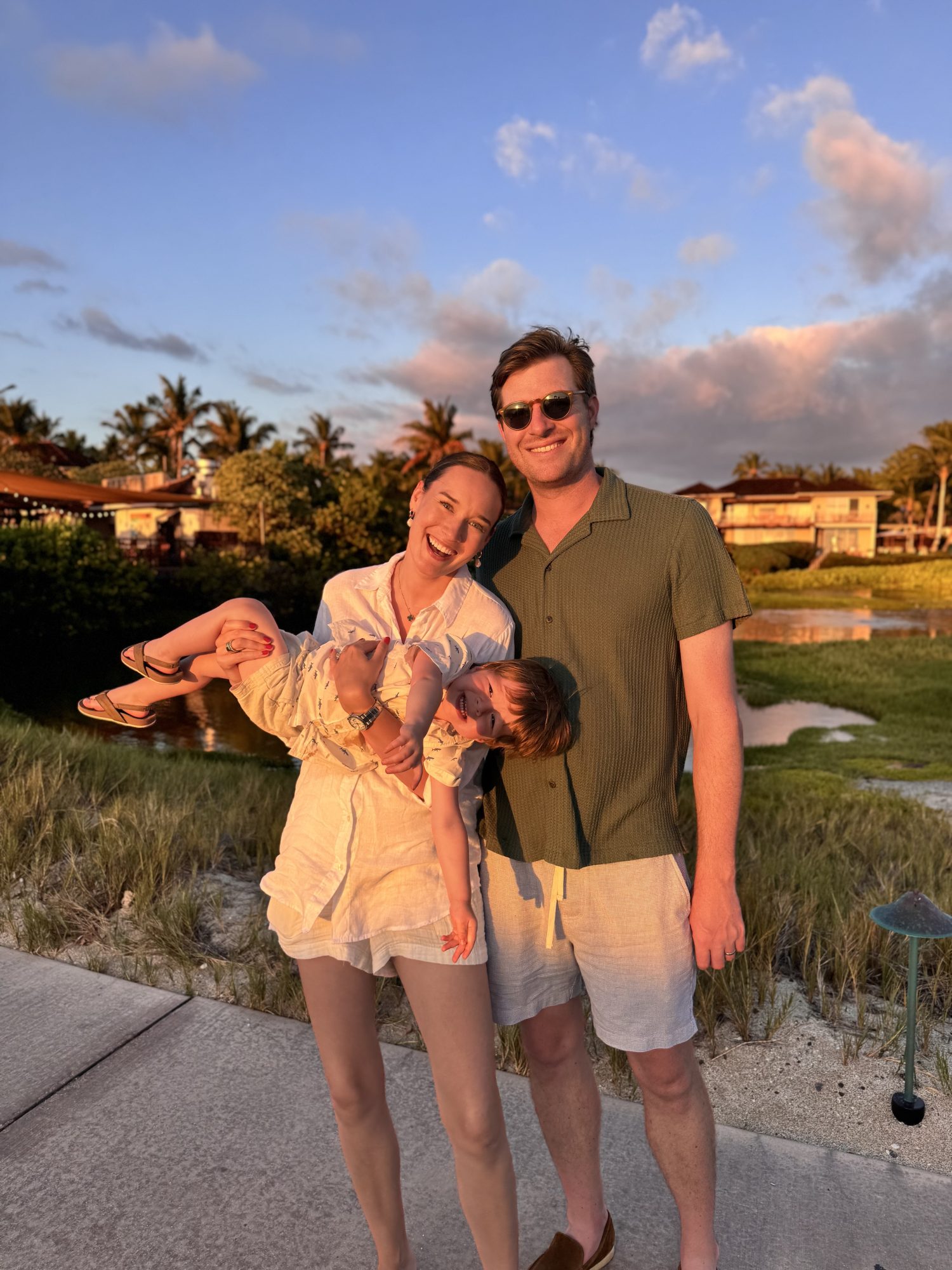 Three people stand outdoors on a paved path at sunset; an adult woman holds a child, who is upside down, while another adult stands next to them, all smiling.